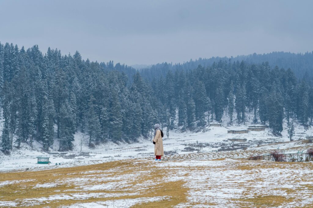 pexels-photo-15432729-15432729 A serene winter scene in Yousmarg featuring a lone figure among snow-covered hills and coniferous trees.